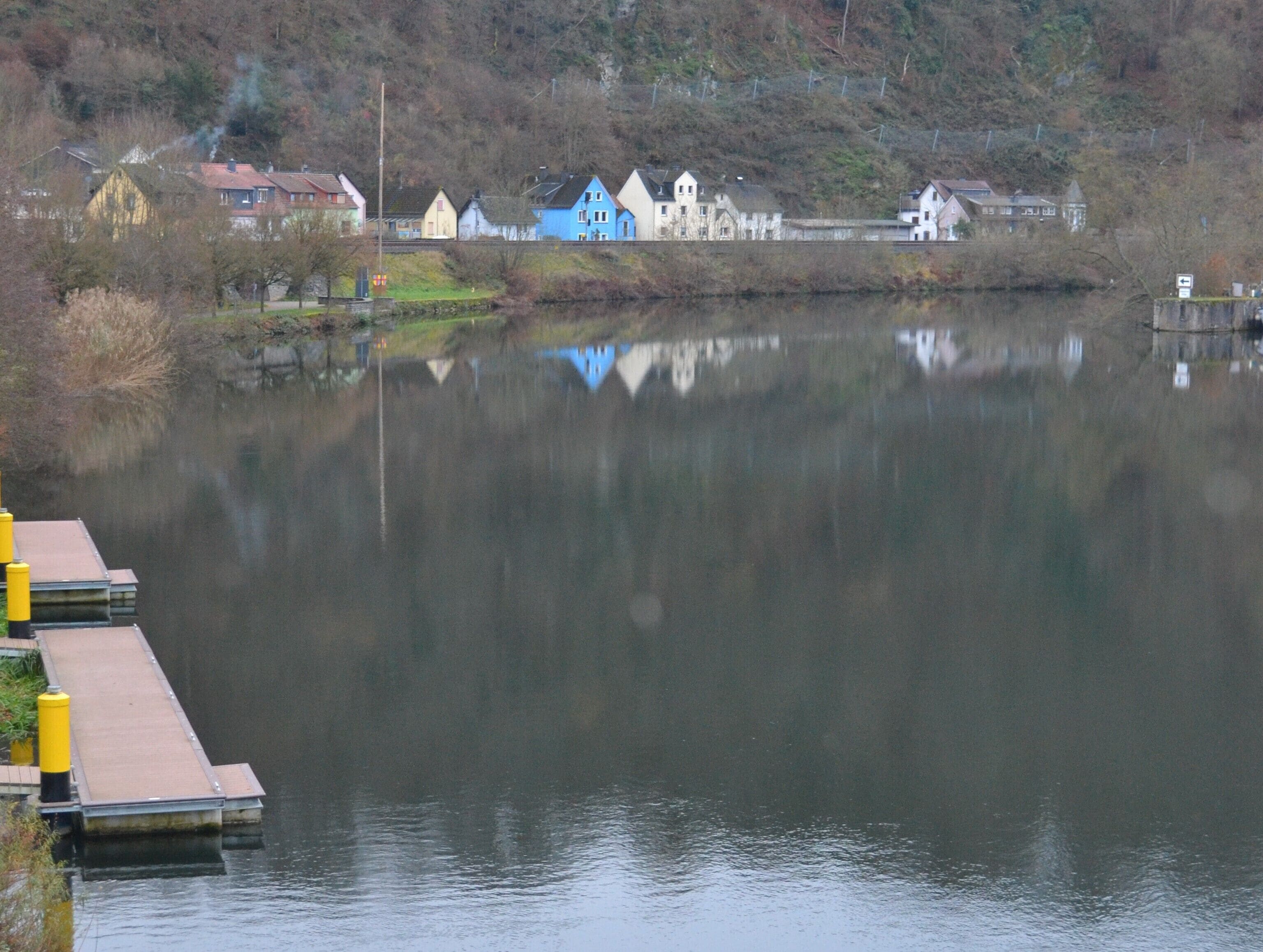 A calm lake with houses reflecting on the water, a dock with yellow bollards, and a hilly backdrop.