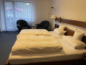 Une chambre d’hôtel avec un grand lit, deux fauteuils, une lampe et une vue à travers de fines rideaux.