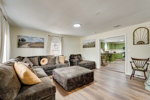 A living room with a sectional sofa, ottoman, and a dining area with a kitchen in the background. - Historic Louisville Steps Away! Work & Play Home (Louisville)