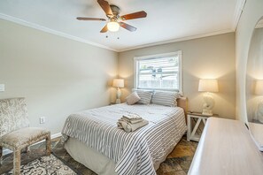 Ein Schlafzimmer mit Bett, Sessel, Nachttisch mit Lampen, Deckenventilator und Fenster mit Jalousien.