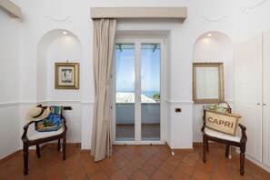 A room with a view through a glass door, a chair with a hat, a bag labeled 'CAPRI', and a framed picture on the wall. - Villa Preziosa - Panoramic Sea View (Capri)