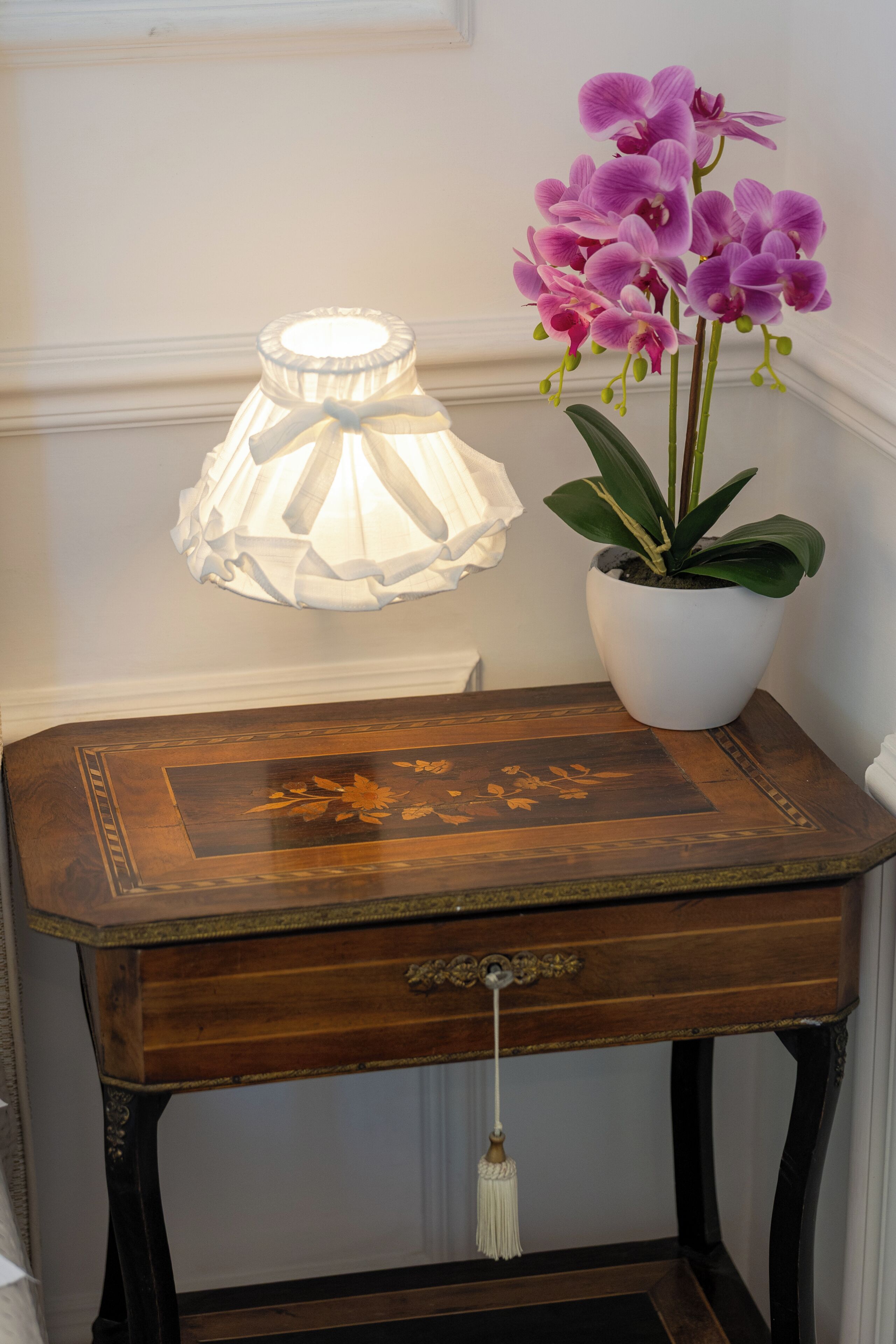 A wooden table with a floral design, a potted plant with purple flowers, and a white lamp.