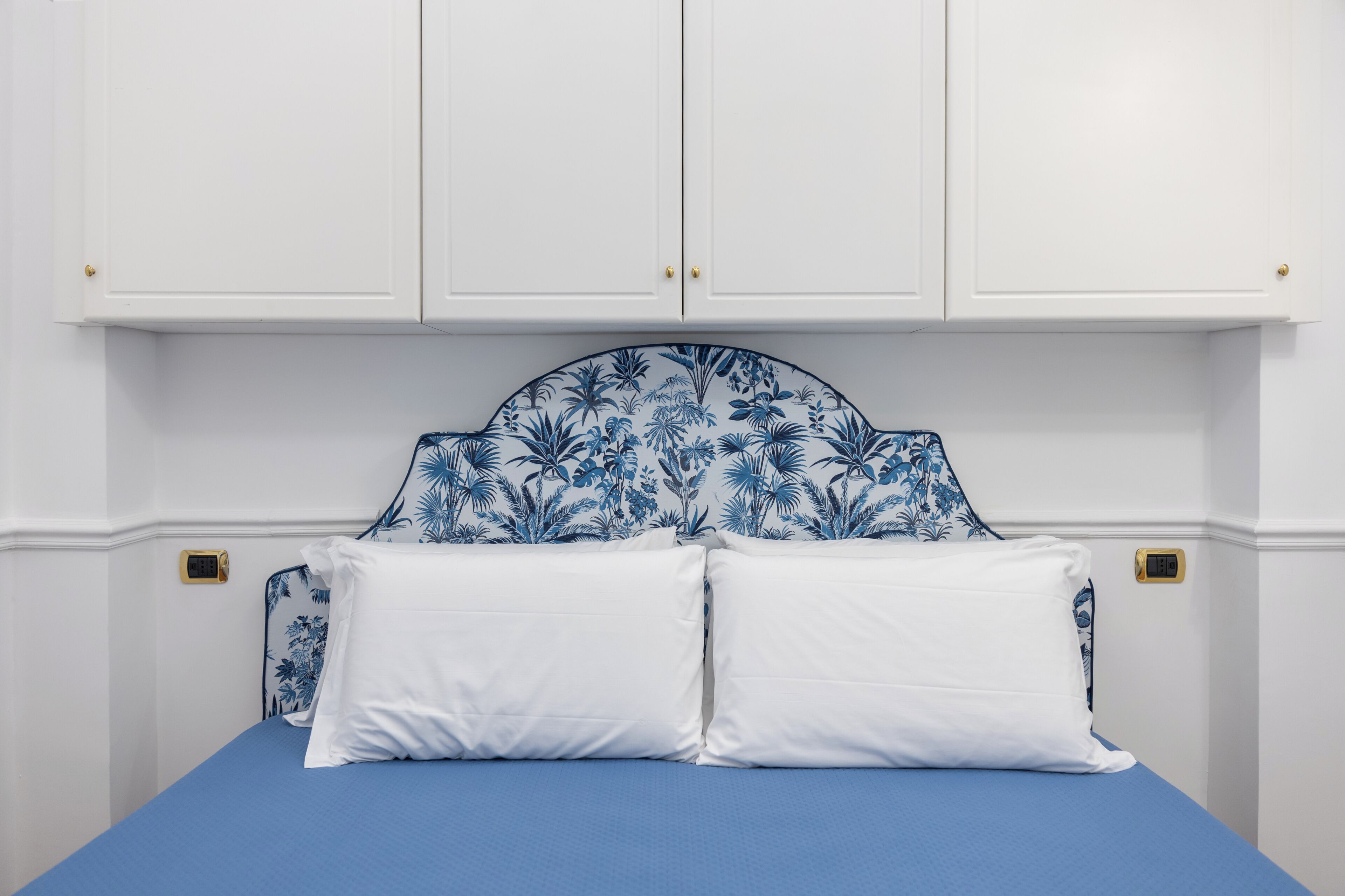 Een slaapkamer met een bed, witte muren en een blauw hoofdeinde met een bloemenpatroon.