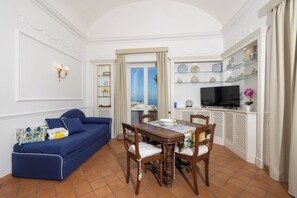 A living room with a blue sofa, wooden dining table, chairs, a TV, and shelves with decorative items. - Villa Preziosa - Panoramic Sea View (Capri)