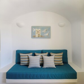 Ein lindgrünes Sofa mit gemusterten Kissen, ein Wandbild mit einem Gebäude und Vögeln sowie eine weiße Wand mit zwei Wandleuchten.