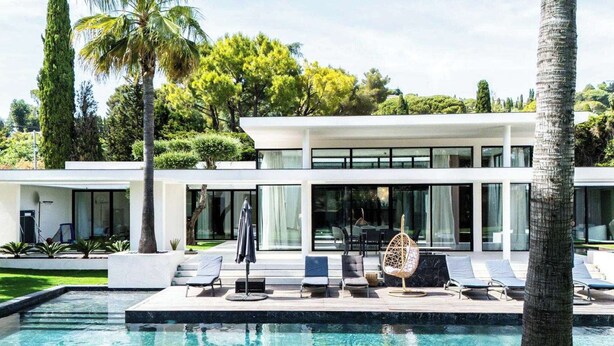 Een modern huis met een zwembad, lounge stoelen en een tropische tuin.