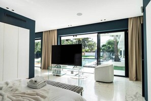 Ein modernes Hotelzimmer mit Bett, TV-Stand aus Glas, einer Sitzecke und Blick auf einen Swimmingpool.