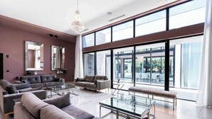 Een moderne woonkamer met grote ramen, een kroonluchter en grijze banken.