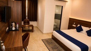 A hotel room with a bed, a desk, a chair, a small table, an air conditioning unit, and a bathroom visible through a glass door. - Hotel Raaso Inn (Jaipur)