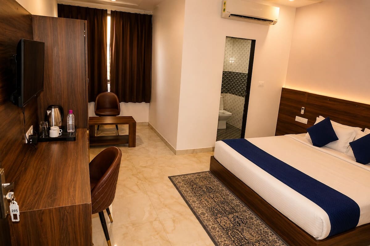 Ein Hotelzimmer mit einem Bett, einem Schreibtisch, einem Stuhl, einem kleinen Tisch, einer Klimaanlage und einem durch eine Glastür einsehbaren Badezimmer.