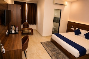 A hotel room with a bed, a desk, a chair, a small table, an air conditioning unit, and a bathroom visible through a glass door. - Hotel Raaso Inn (Jaipur)