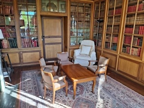Een bibliotheek met houten boekenkasten, een witte fauteuil, een houten tafel en vloerkleden met patronen.