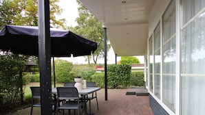 A covered patio with a table and chairs, a potted plant, and a view of a building with large windows. - Holiday Home in Brunssum With Terrace (Brunssum)