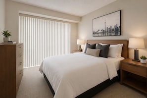 A hotel room with a large bed, two bedside tables with lamps, a cityscape painting, a window with blinds, and a wooden dresser. - Solara & Slate Hotel (Toronto)