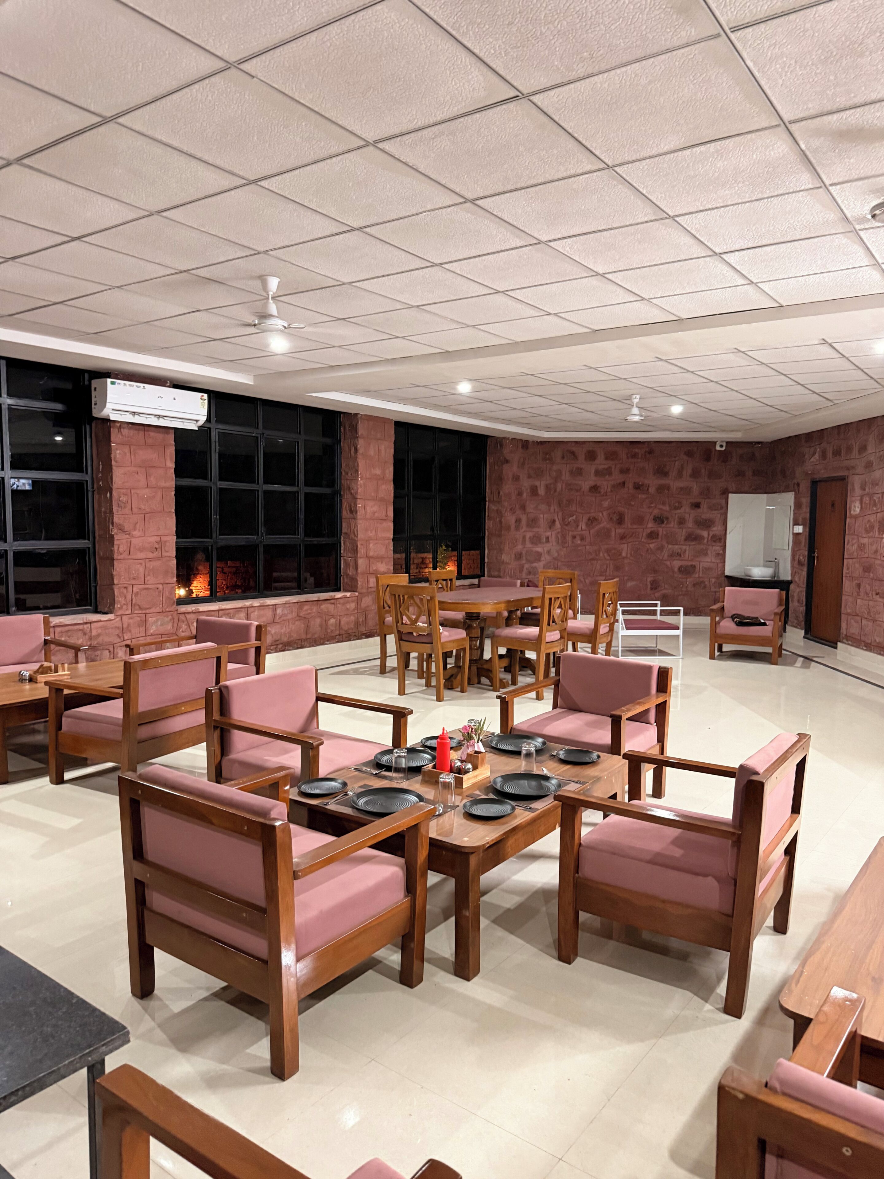 A spacious indoor area with multiple wooden tables and chairs, a ceiling fan, and a large window with a view of a fire.