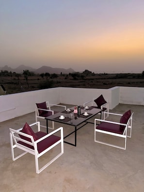 Une terrasse sur le toit avec une table en verre, des chaises blanches avec des coussins rouges et une vue sur les montagnes lointaines.
