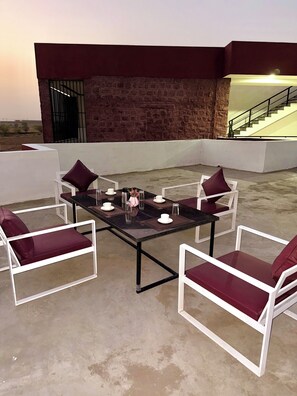 A rooftop patio with a table set for four, surrounded by white chairs with red cushions, a brick wall, and a staircase leading to an upper level.