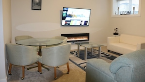 A living room with a glass table, two patterned chairs, a white sofa, a flat-screen TV mounted on the wall, a fireplace, and a window with the word 'DREAM' on it.