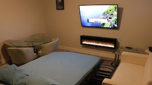 A hotel room with a bed, a glass table, a sofa, a TV mounted on the wall, and a fireplace.