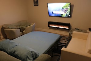 A hotel room with a bed, a glass table, a sofa, a TV mounted on the wall, and a fireplace. - Lovely Guest Suite in Calgary (Calgary)