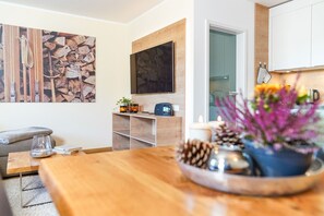 Ein modernes Wohnzimmer mit einem Holztisch, einem an der Wand montierten Fernseher und einem durchgehenden Gang sichtbaren Küchenbereich.