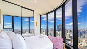 A hotel room with a large bed, two bedside tables, and a view of a cityscape through large windows. - SKY HIGH Apartment (Melbourne)