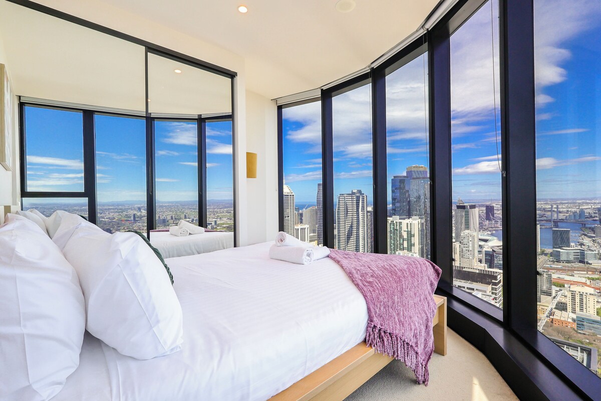 Ein Hotelzimmer mit einem großen Bett, zwei Nachttischen und Blick auf eine Stadtkulisse durch große Fenster.