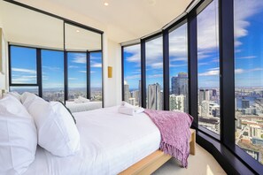 Ein Hotelzimmer mit einem großen Bett, zwei Nachttischen und Blick auf eine Stadtkulisse durch große Fenster.