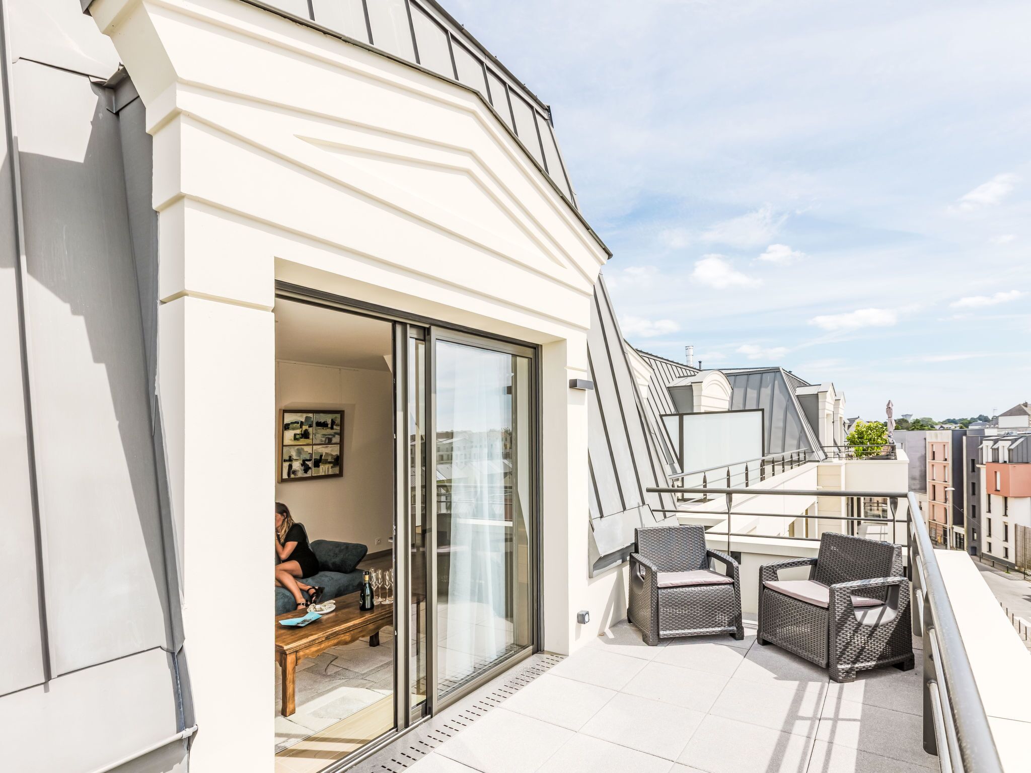 Un bâtiment moderne doté d’un balcon, d’une entrée avec porte vitrée et d’un espace extérieur aménagé pour s’asseoir.