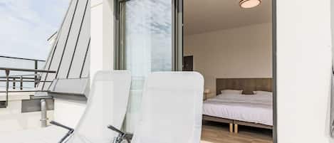 A modern hotel room with a large sliding glass door leading to a balcony with two white lounge chairs.