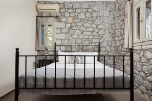 A bed with a metal frame, a stone wall, a window with curtains, and an air conditioning unit. - Prosilion Stone House (Kotronas)