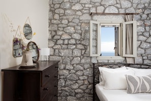 A room with a stone wall, a bed with white bedding, a dresser, a mirror, and a window with curtains.