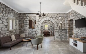 A stone-walled interior with a sofa, armchair, dining table, and TV. - Prosilion Stone House (Kotronas)