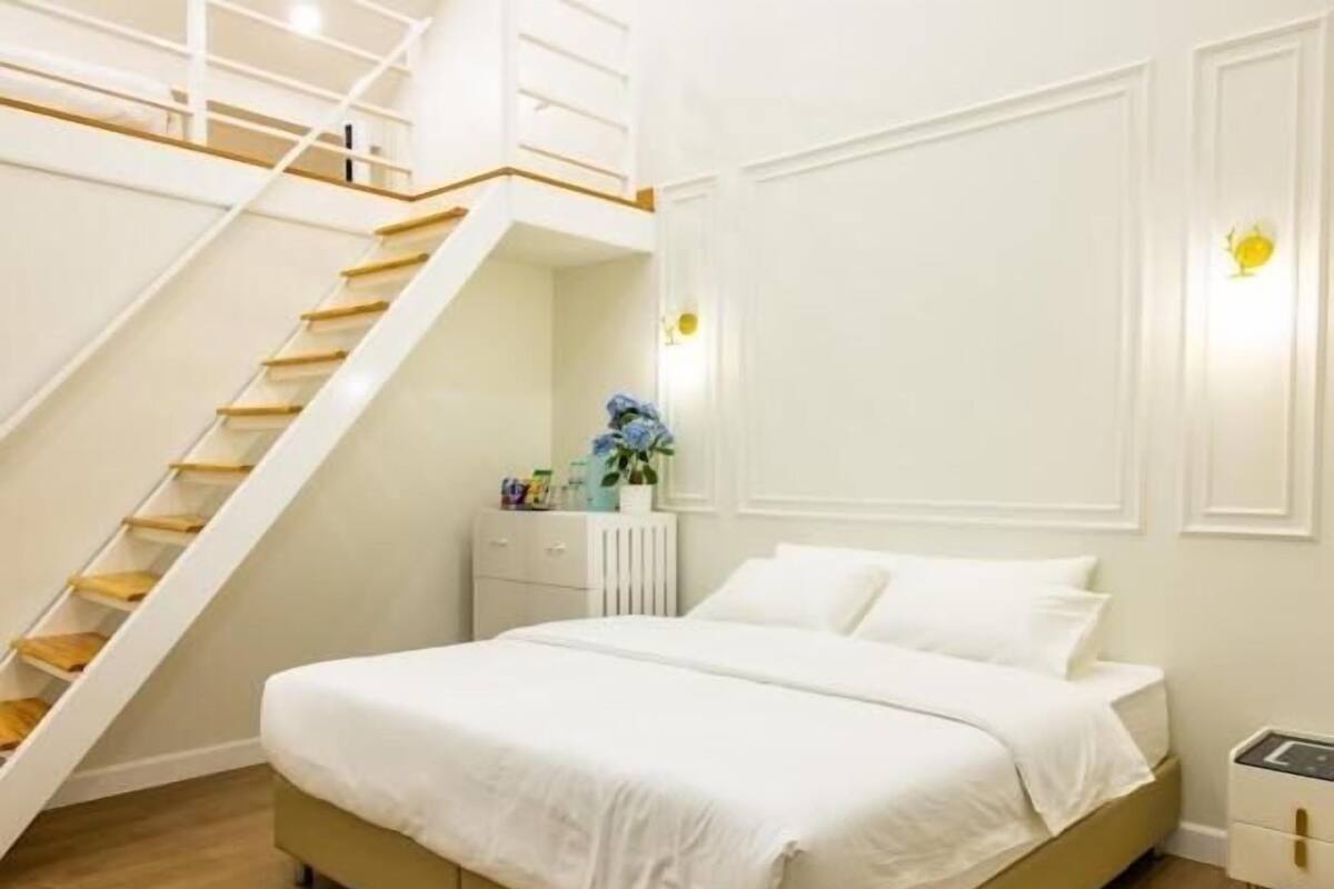 Un dormitorio con cama, una mesita de noche blanca, un aparador y una pared blanca con interruptor de luz, además de una escalera.