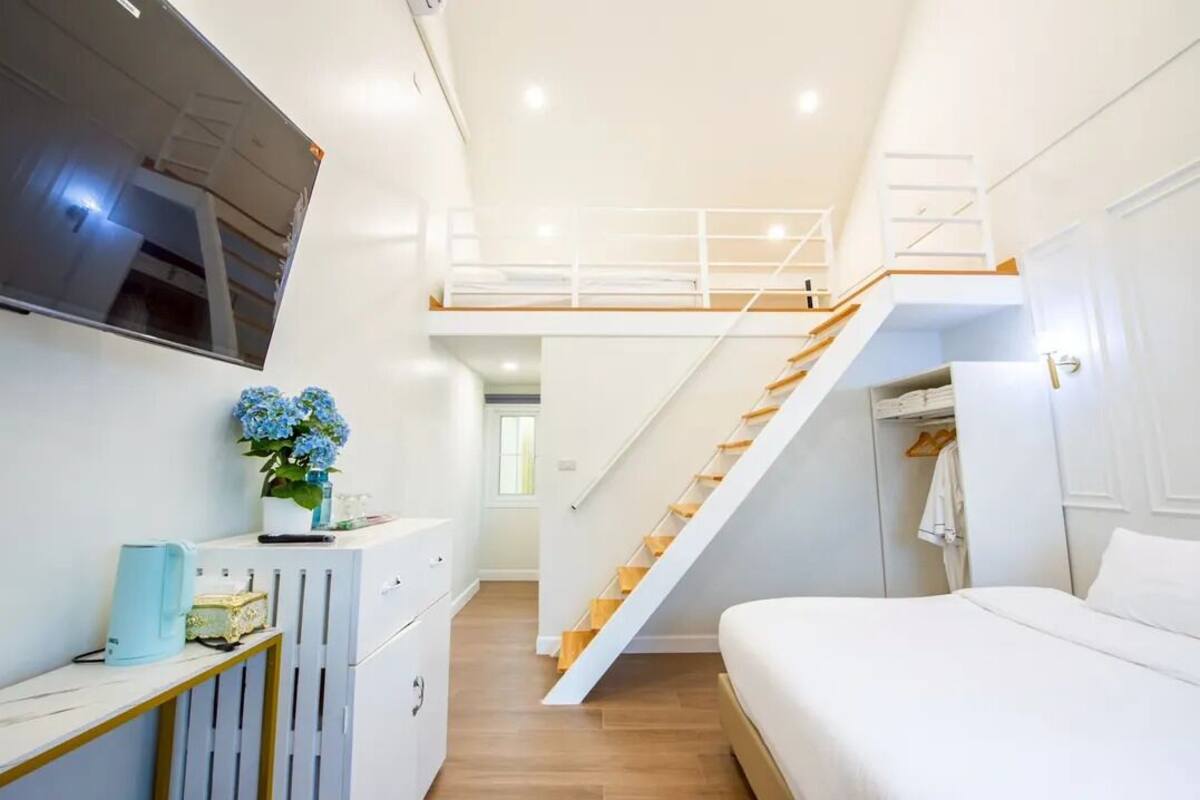 Un dormitorio con cama, televisor montado en la pared, escalera y un pequeño escritorio con planta.