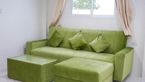 Un sofá verde con otomana y cojines a juego, una mesa de centro de vidrio y una ventana con cortinas.