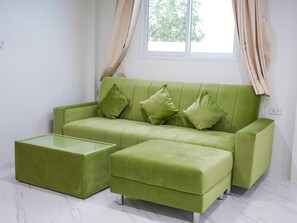 A green sofa with matching ottoman and cushions, a glass coffee table, and a window with curtains.