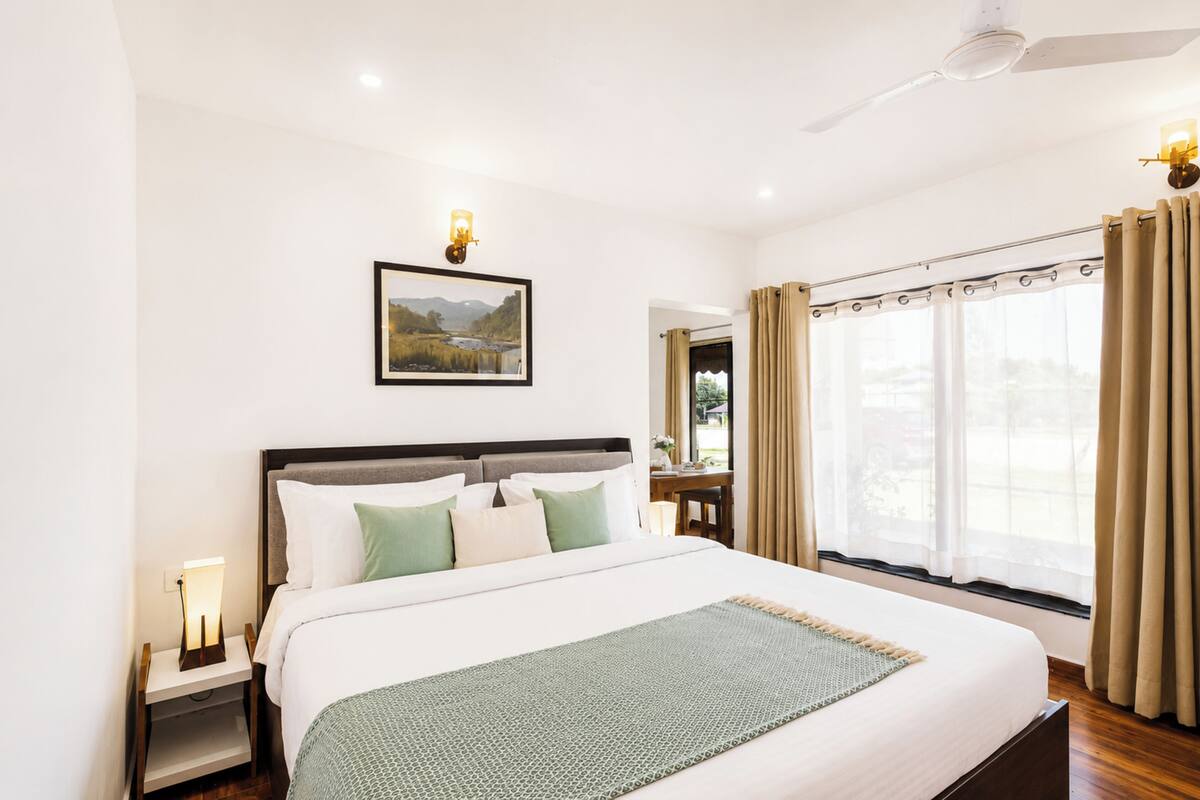 A neatly arranged hotel room with a large bed, a bedside table with a lamp, a ceiling fan, a window with curtains, and a framed picture on the wall.