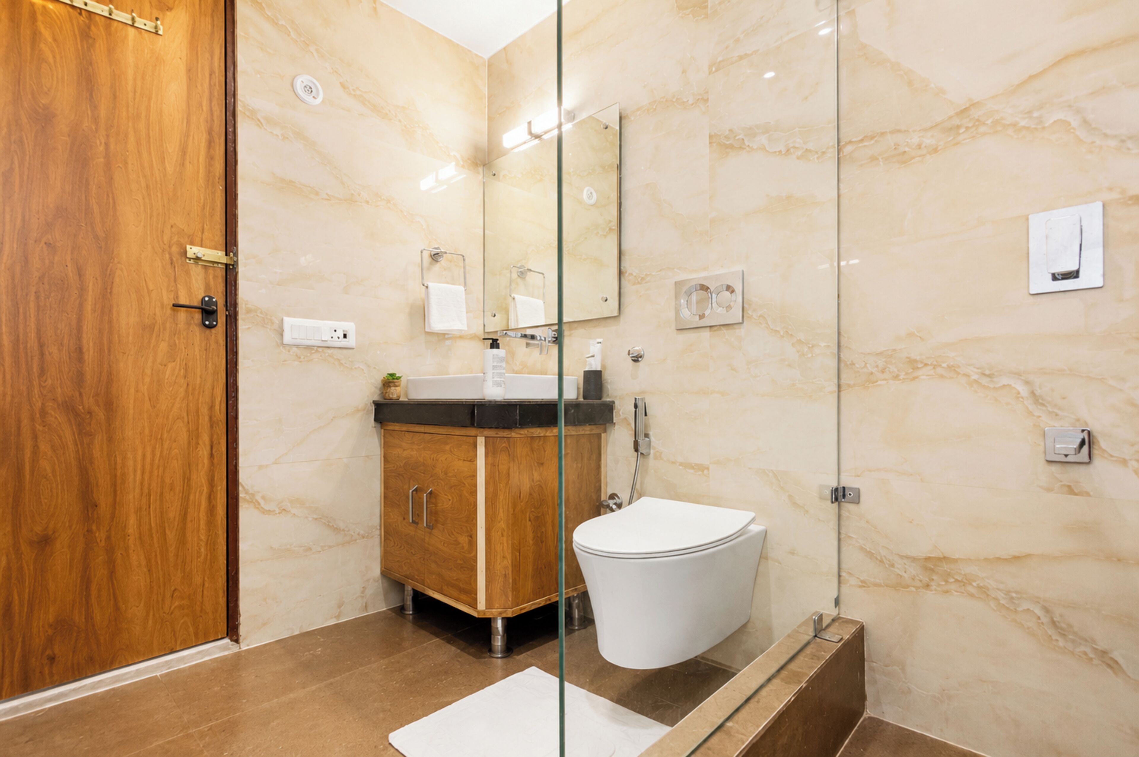 A modern bathroom with a wooden door, marble walls, a glass shower enclosure, a sink with a mirror, and a toilet.