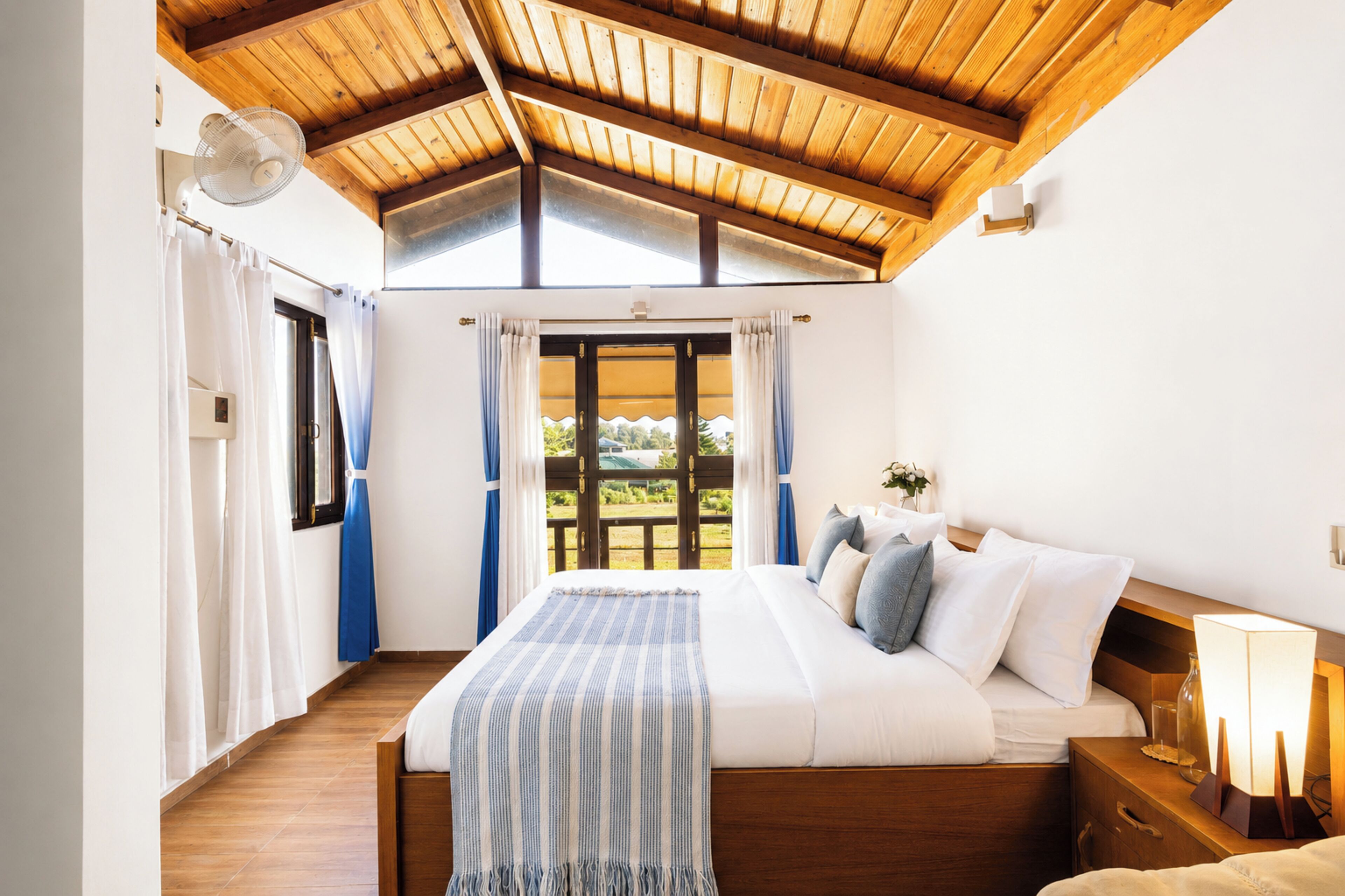 A bedroom with a wooden ceiling, a bed with white bedding, a bedside table with a lamp, and a view through a window.