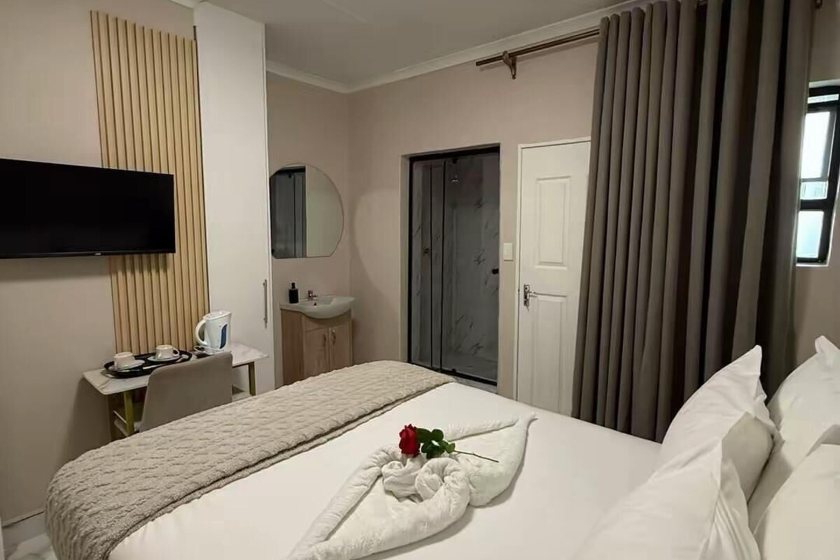 A hotel room with a bed, a TV, a desk with a kettle, a mirror, and a door.