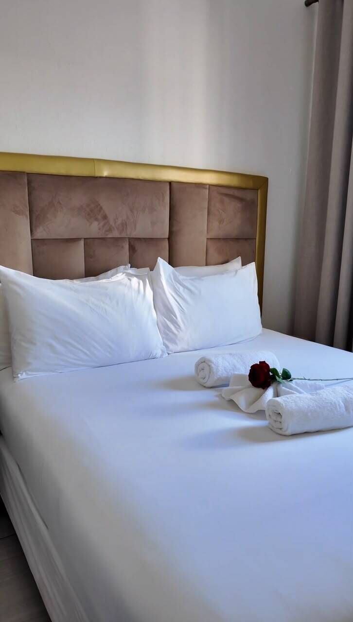 A neatly made bed with white linens and pillows, a gold headboard, and a single red rose on the bed.