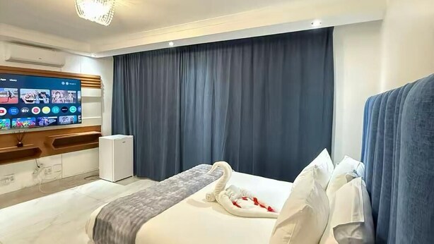 A hotel room with a bed, a TV mounted on the wall, a bedside table with a lamp, and a window with curtains.