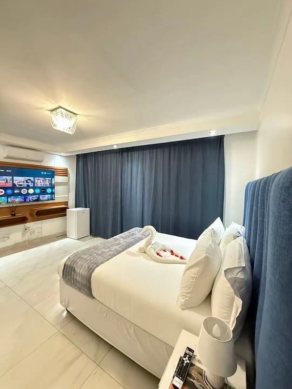 A hotel room with a bed, a TV mounted on the wall, a bedside table with a lamp, and a window with curtains.