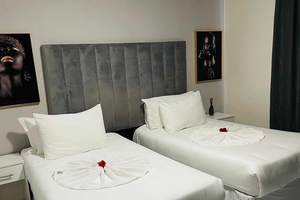 Quarto de hotel com duas camas, cabeceira cinzenta, roupa de cama branca, tapete listrado e obras de arte nas paredes.