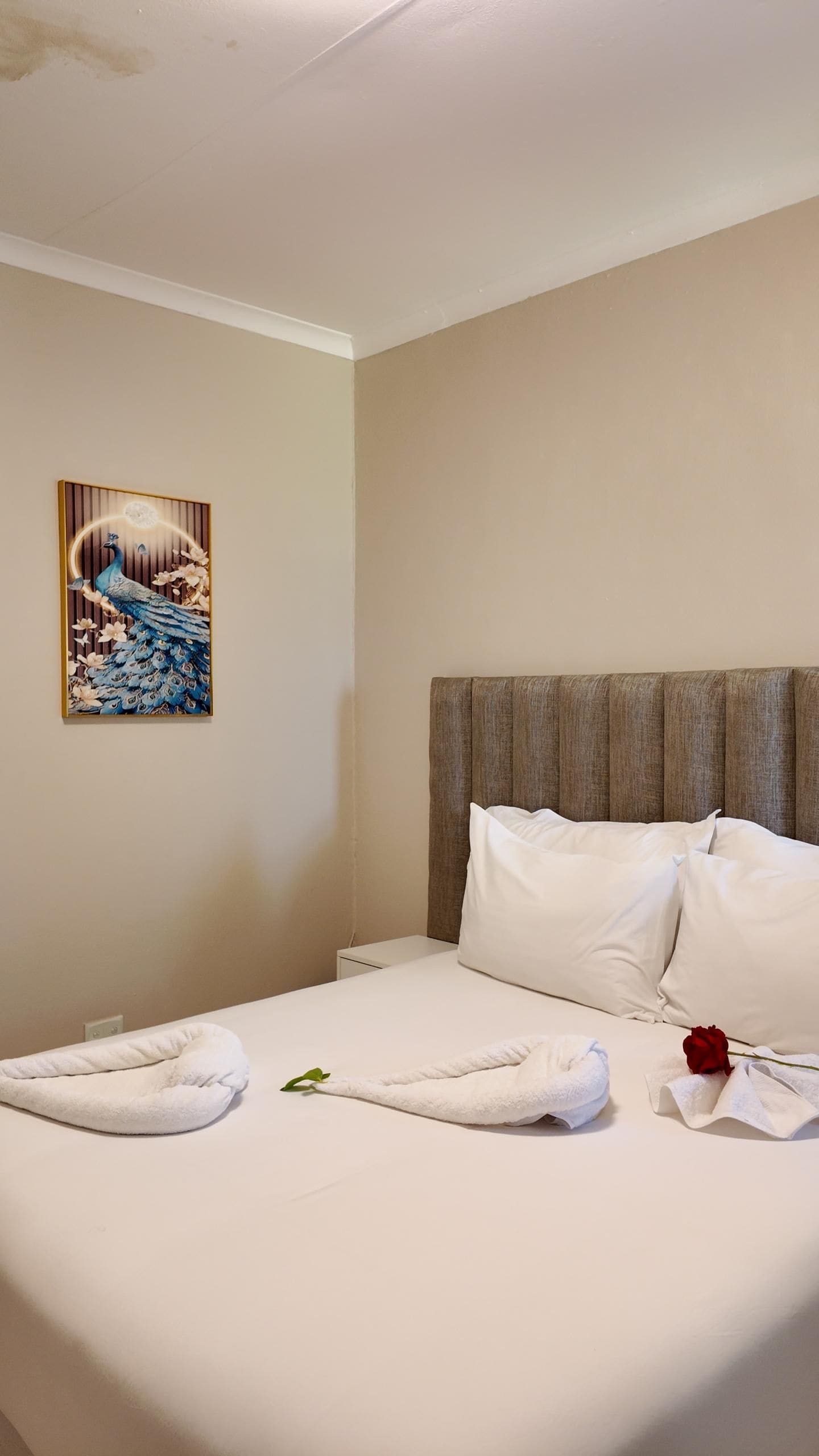 Hotellihuone, jossa on sänky, tyynyt, sängynpääty, seinälle ripustettu taulu ja ruusukuviollinen pyyhe.