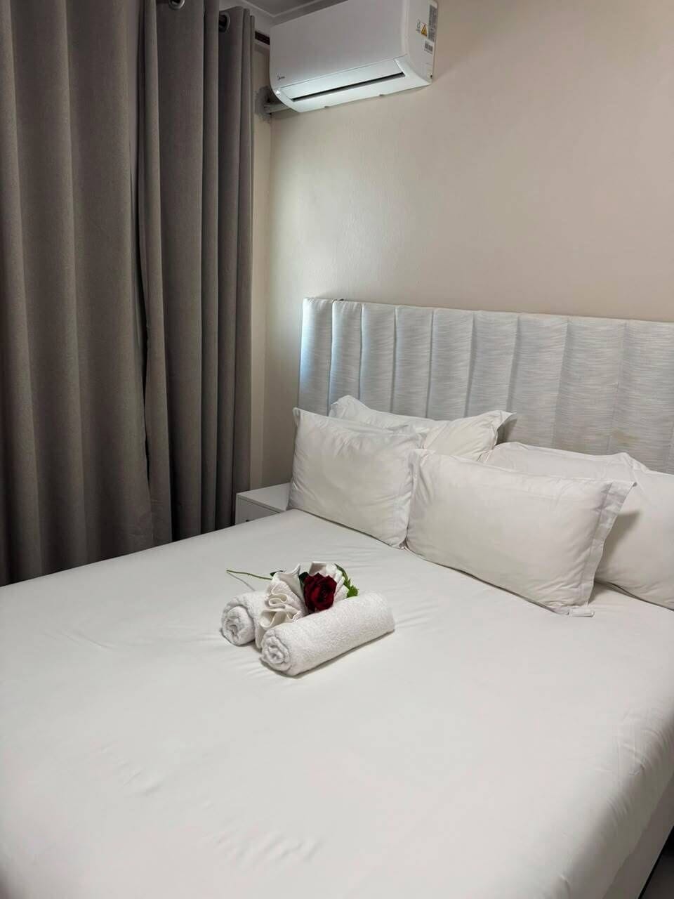 A hotel room with a bed, pillows, a rolled towel, and an air conditioning unit.
