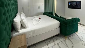 A modern bedroom with a green tufted headboard, a white bed, a green sofa, a wooden nightstand, and a TV mounted on the wall.