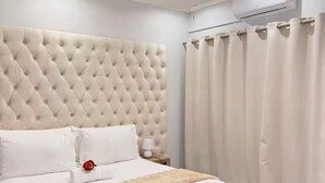A bedroom with a tufted headboard, a bed with white linens, a bedside table with a lamp, and a large window with curtains.