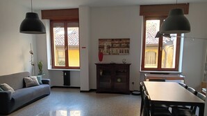 Sala de estar com sofá, mesa de jantar, cadeiras, radiador e janela com vista para edifícios.